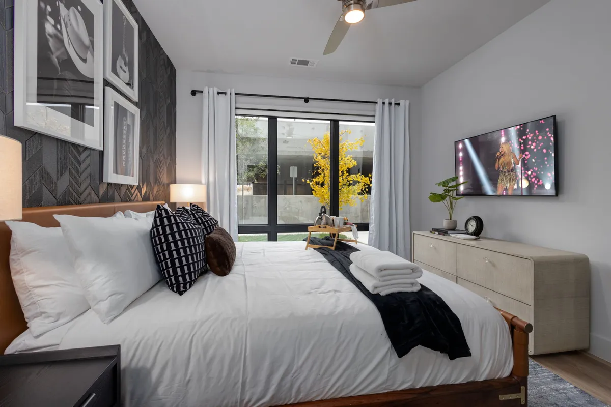 This image features a bedroom with white linens, framed art on the walls, large windows letting in natural light, a television, and a green plant. The space exudes a calming and cozy atmosphere, with modern décor and thoughtful accents enhancing comfort.