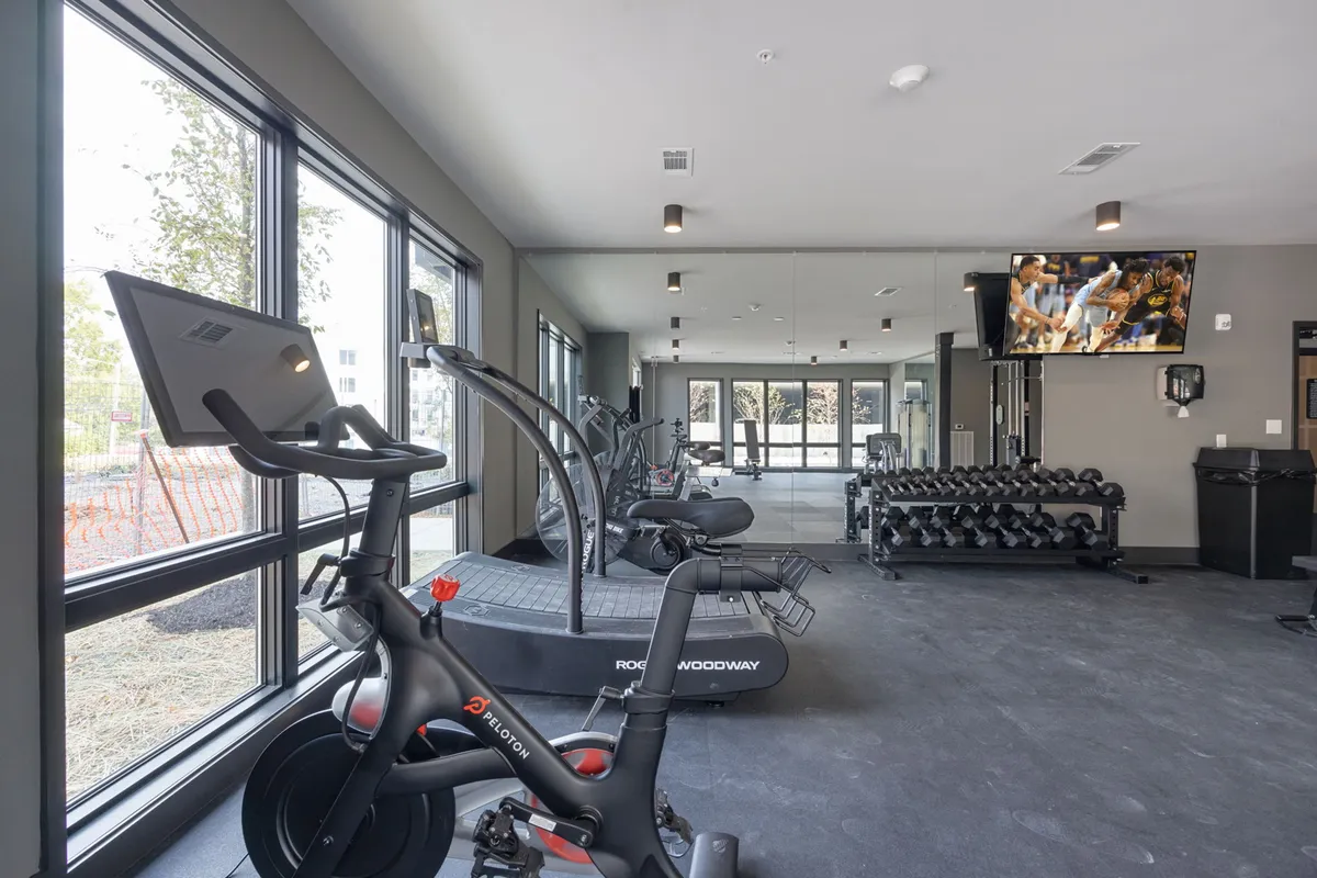 A bright interior gym with a Peloton bike, treadmill, dumbbell rack, and large windows letting in natural light. The space is modern and energetic, ideal for maintaining fitness routines just steps from the condos.