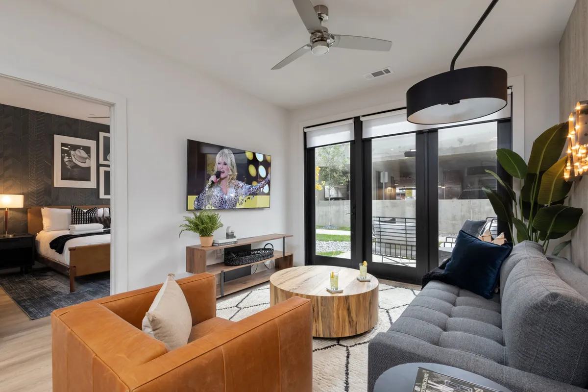 A living room featuring a comfortable seating arrangement, a television displaying a performance, and expansive windows letting in plenty of sunlight. The space feels warm and inviting, making it perfect for movie nights or relaxed group interactions.