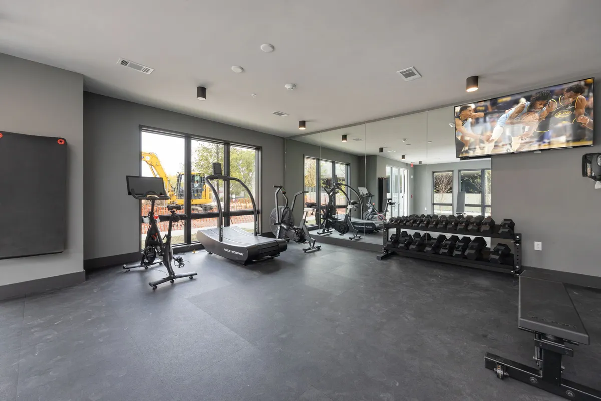 This image depicts a well-equipped on-site fitness center, complete with a treadmill, exercise bike, dumbbell rack, large windows, and a television. The space is filled with natural light, supporting both personal and group workouts.