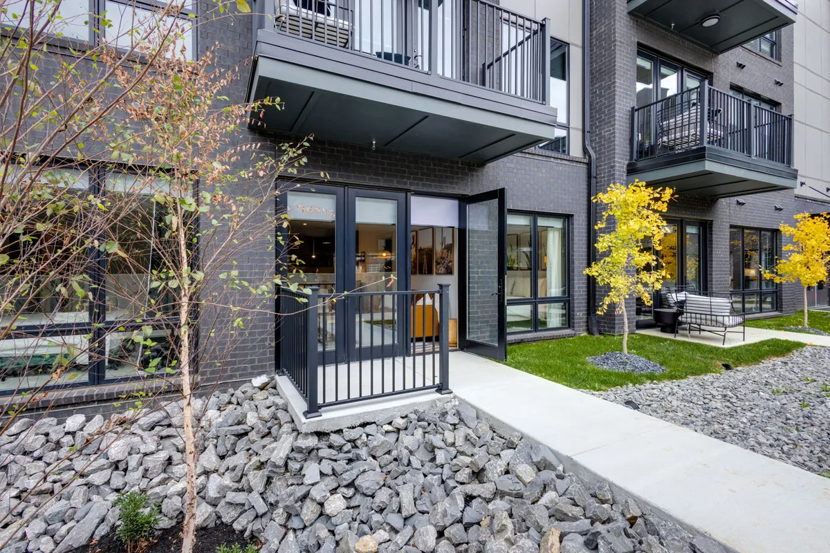 The image shows the entrance to a contemporary condo building, featuring modern design elements, decorative stones, yellow trees, and balconies. The atmosphere is welcoming, with spaces for outdoor relaxation and socializing just outside the door.