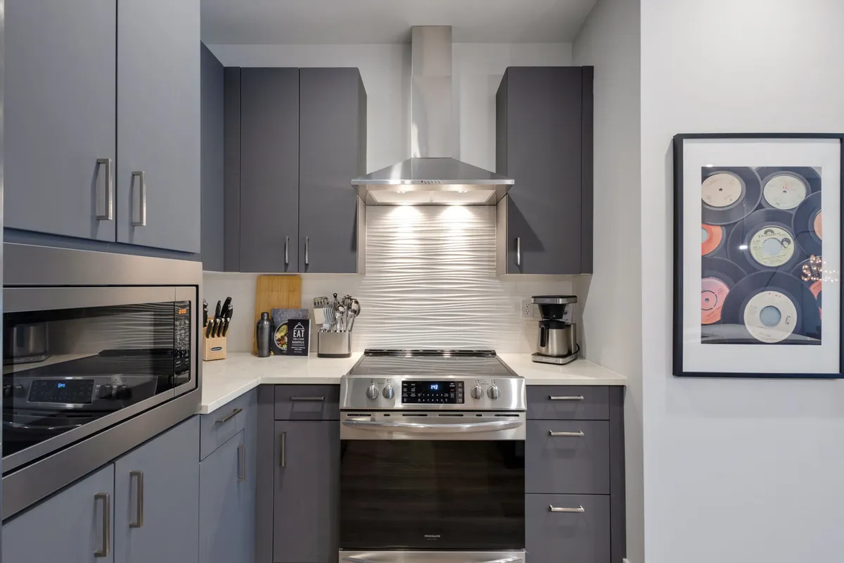 A stylish kitchen with stainless steel appliances, textured backsplash, and sleek gray cabinetry. Contemporary art adorns the wall, and the countertop is uncluttered, suggesting a high-functioning, practical cooking environment ideal for shared feasts.