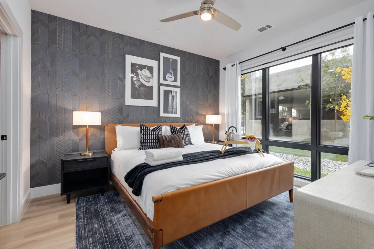 A stylish bedroom with a bed featuring a leather headboard, framed artwork on the wall, and large windows that flood the room with natural light. The overall vibe is comfortable and contemporary, ideal for unwinding.